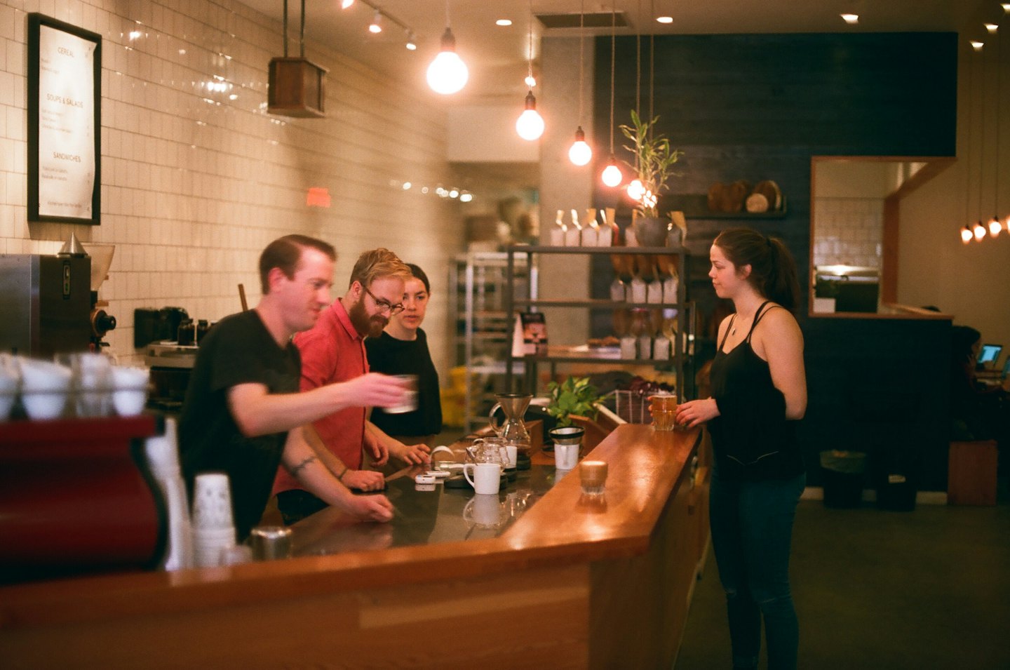 Barista obsluhuje zákazníky u pultu útulně osvětlené kavárny.